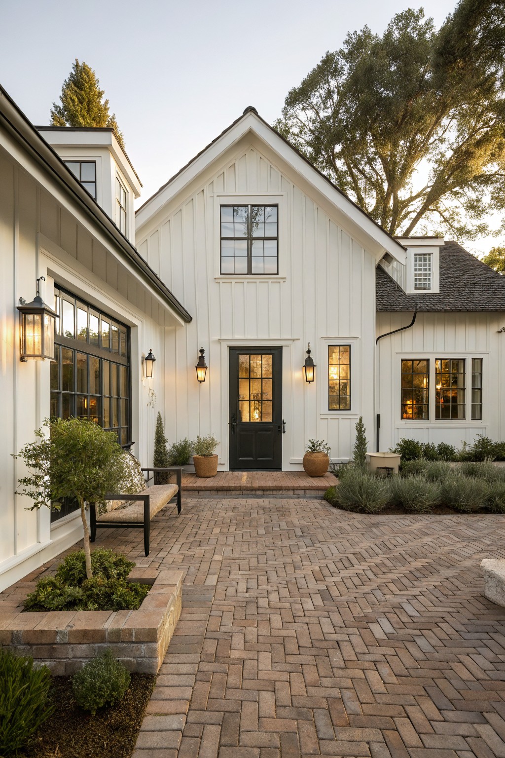 White board-and-batten single-story farmhouse exterior with black front door and windows, flanked by lanterns, brick paver entry landing and path, bench with cushions, potted plants, low shrubs, and trees in the background.