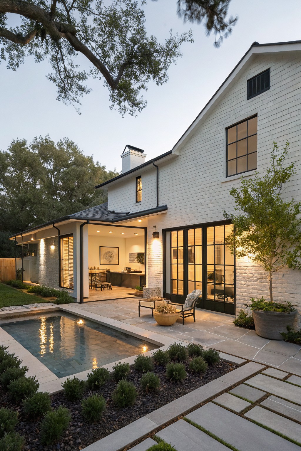 Rear exterior of a single-story white shiplap house with black-framed glass doors opening to a covered patio area featuring wicker seating, a round planter, and an adjacent narrow rectangular pool with paver surround and low shrubs.