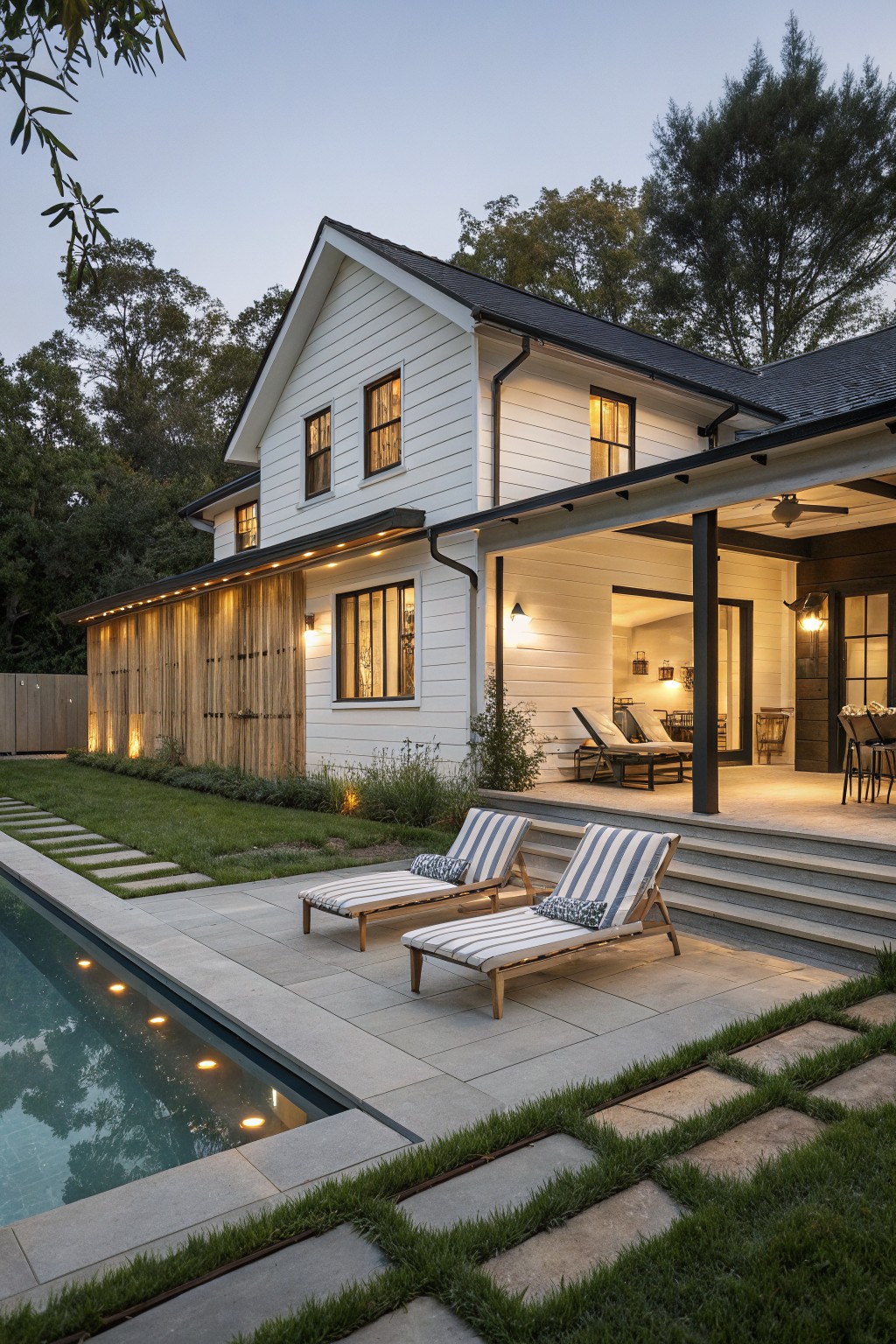 Modern white farmhouse with pool, patio, and striped lounge chairs at dusk.