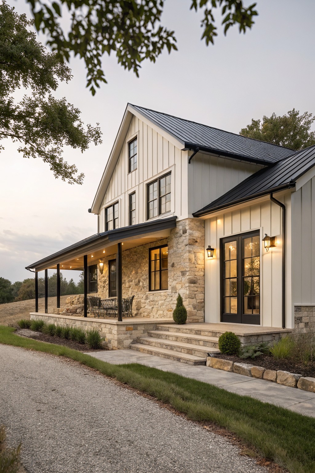 Single-story modern farmhouse exterior featuring white board-and-batten siding, black metal roof, beige stone foundation and porch accents, covered front porch with black-trimmed doors and lanterns, stone steps, gravel driveway, and surrounding lawn and trees.