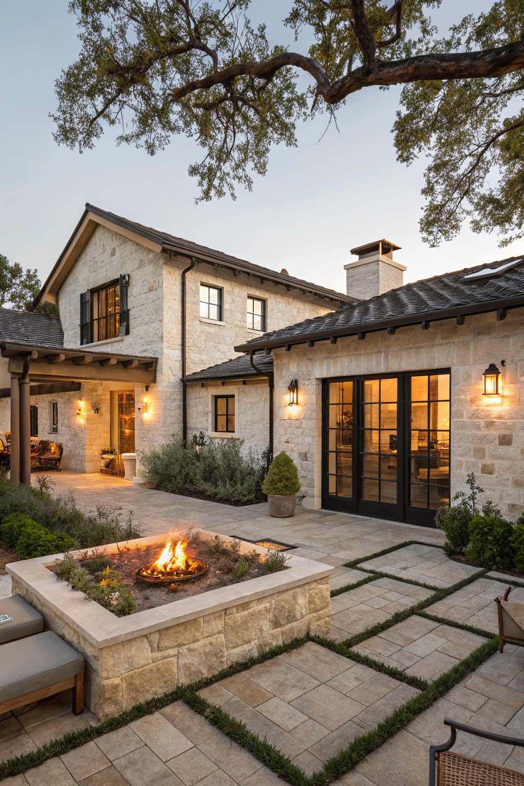 Stone house exterior with covered porch, black-framed glass doors, lantern lights, and adjacent patio with rectangular built-in fire pit containing flames, paver tiles with grass inlays, and cushioned lounge seating.