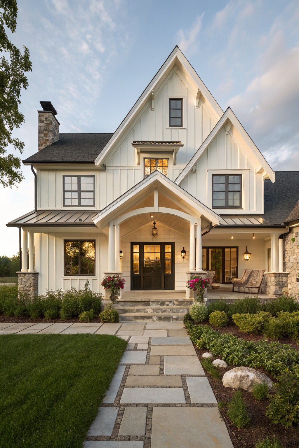 White board-and-batten sided modern farmhouse house exterior with black windows, gabled dark shingle roof, covered front porch supported by stone posts, stone chimney, flower planters, and flagstone pathway through lawn and landscaping.
