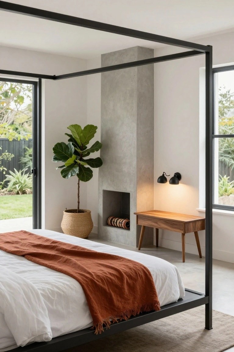 A bedroom with a black metal canopy bed draped in white sheets and an orange throw, next to a concrete fireplace wall, wooden side table, fiddle leaf fig in a woven basket, and large windows to a backyard garden.