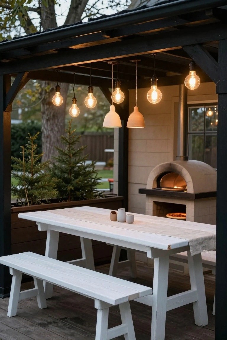 Covered wooden pergola with multiple hanging bulb lights over a white trestle table and benches on a deck, next to a brick pizza oven and potted small trees.