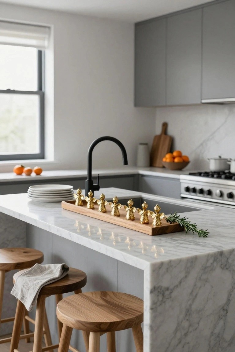 Modern kitchen island with marble countertop holding a wooden tray of gold angel Christmas ornaments and rosemary sprigs, flanked by wooden stools, black faucet, and gray cabinets.