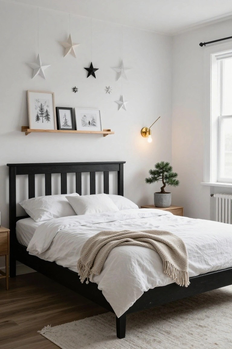 Cozy bedroom corner with black wooden bed frame, white bedding and throw, hanging white and black paper stars from ceiling, wooden floating shelf holding three framed black-and-white winter tree prints, gold wall lamp lit, gray pot bonsai on wood nightstand, white walls, hardwood floor, and seagrass rug.