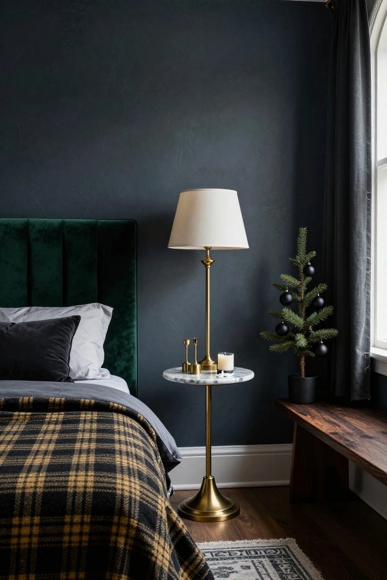 Bedroom corner with emerald green velvet tufted headboard, gray bedding under black-and-gold plaid blanket, brass floor lamp and side table holding candles next to small potted Christmas tree with black ball ornaments, navy walls, wood bench, and window with dark curtains.