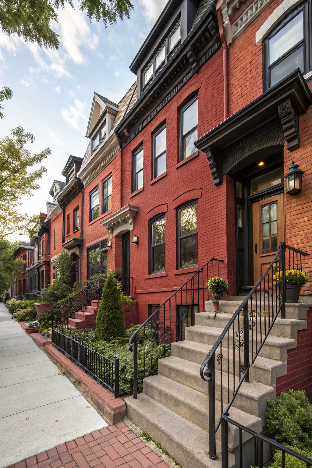 Row of three-story red brick townhouses with black window and door trim, mansard roofs, front stoops with black iron railings and potted plants along a sidewalk.