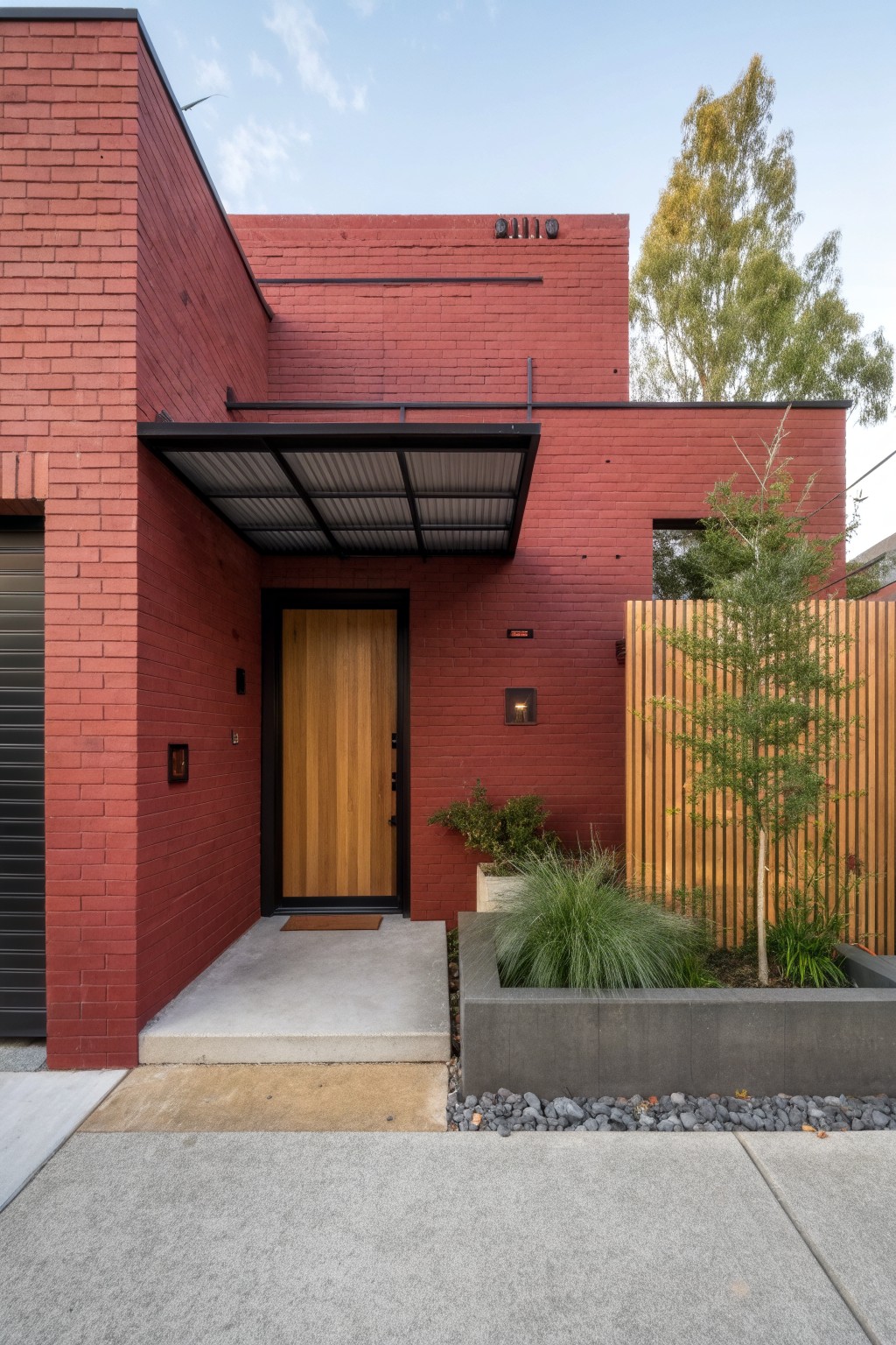 Red brick house exterior with corrugated metal awning over wooden front door and garage, concrete path, and front landscaping including grasses in raised bed and a small tree by wooden fence.
