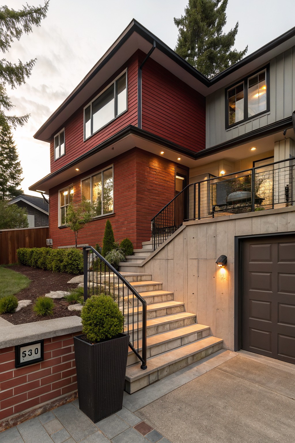 Two-story house exterior with red brick painted lower facade and garage, light gray siding on upper level, concrete stairs with black railings leading to entrance, and surrounding landscaping with shrubs and trees.