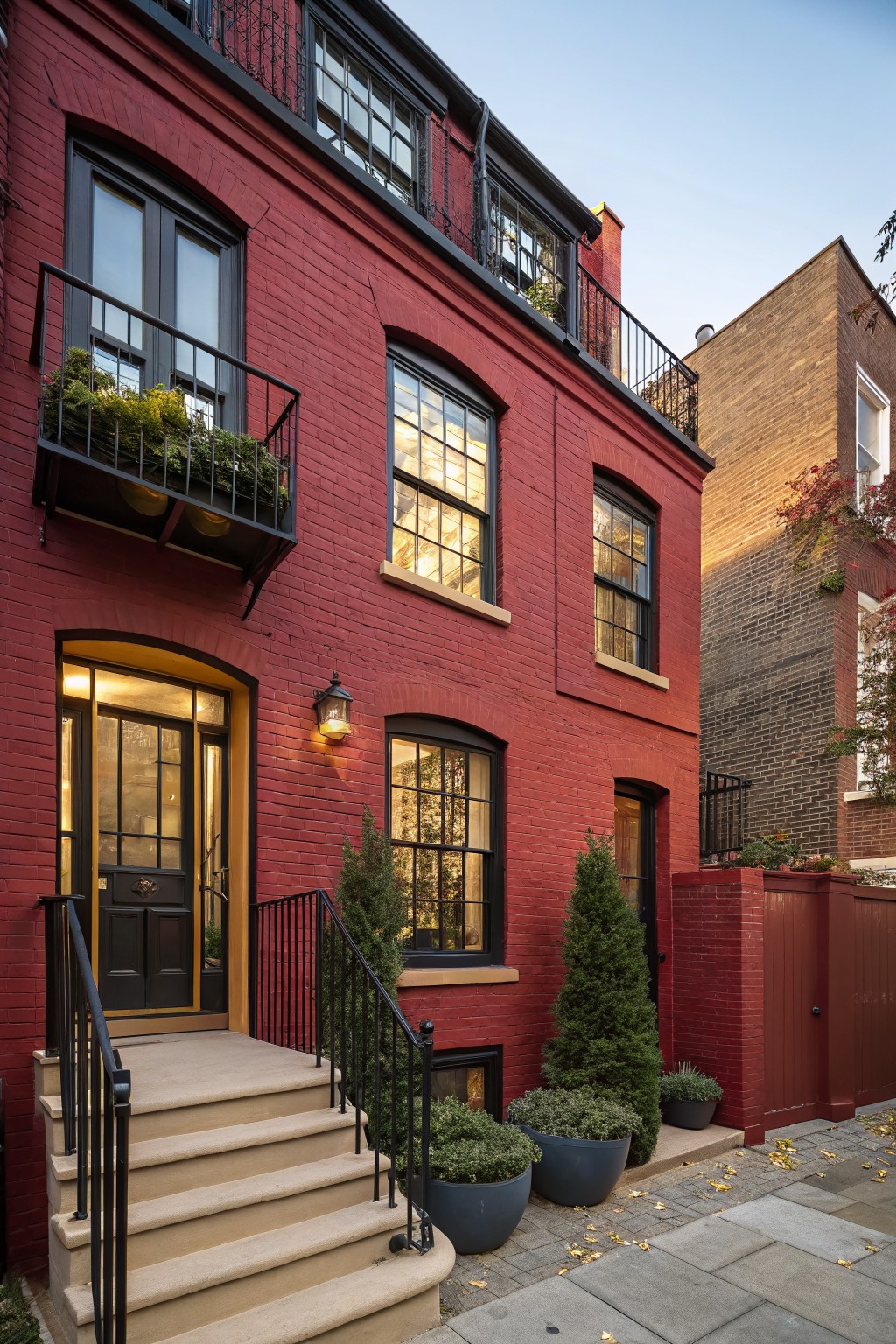 Three-story red brick townhouse with black window frames, wrought iron balconies holding planters, arched yellow entry surround with black door and lanterns, flanked by potted evergreens and shrubs, stone steps to sidewalk, and adjacent tan brick building at dusk.