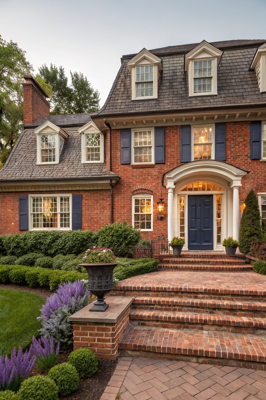 Two-story red brick house exterior featuring dormer windows, blue shutters, white columned arched portico with dark blue door, brick steps, urn planter, and formal shrubs in the front yard.