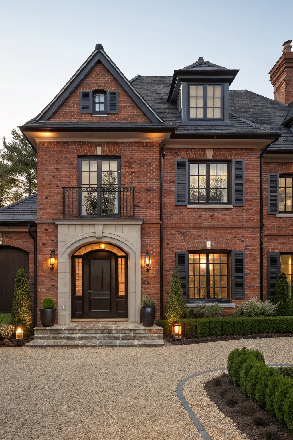 Two-story red brick house exterior featuring a centered arched stone entryway with dark wood door, flanked by lanterns, black-framed windows, and evergreen shrubs along a gravel driveway.
