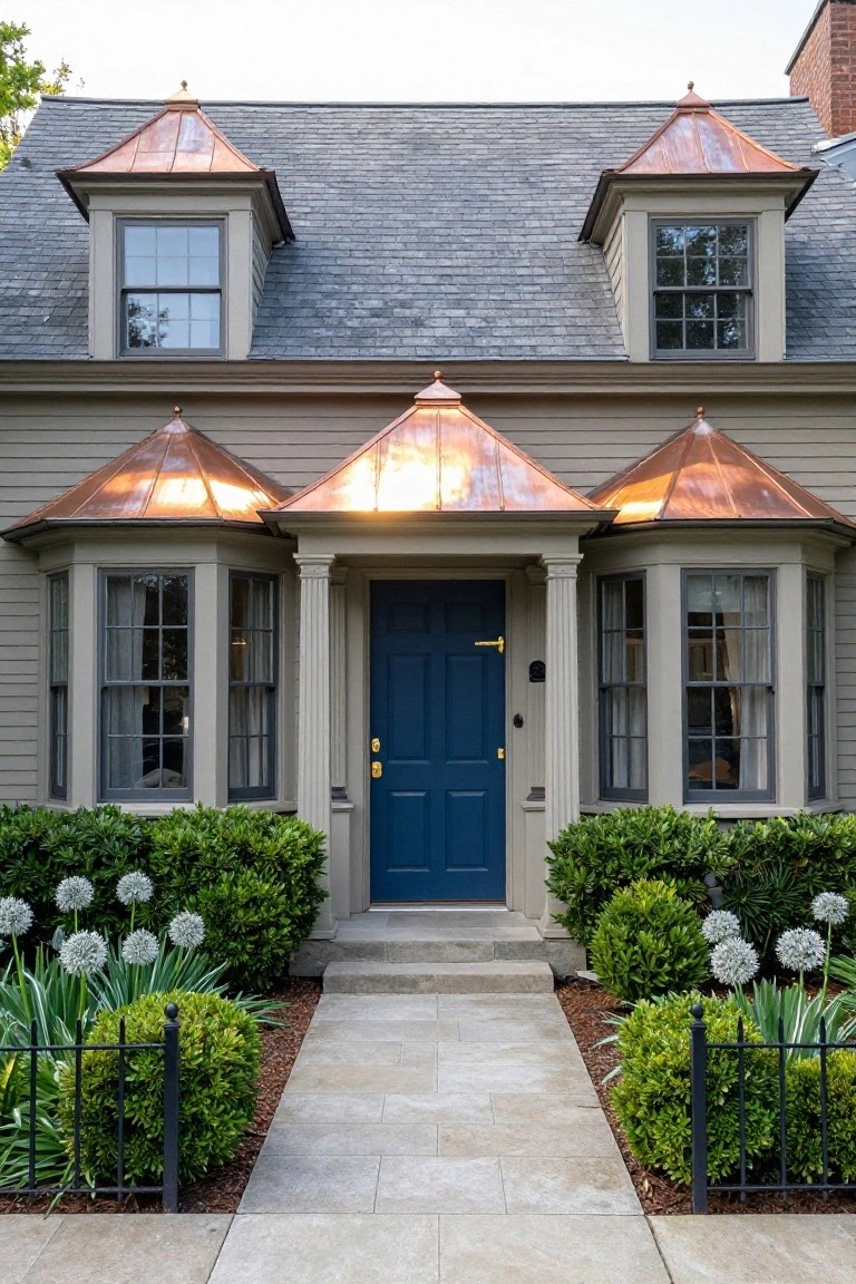 Gray shingled colonial house exterior with copper roofs on two dormers and entry porch, flanked by bay windows, navy blue front door, boxwood shrubs, and stone walkway.