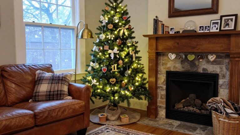 Cozy living room with a lit Christmas tree decorated with wooden heart and initial ornaments, glass balls, and ribbons, beside a leather armchair, bookshelves, and stone fireplace.