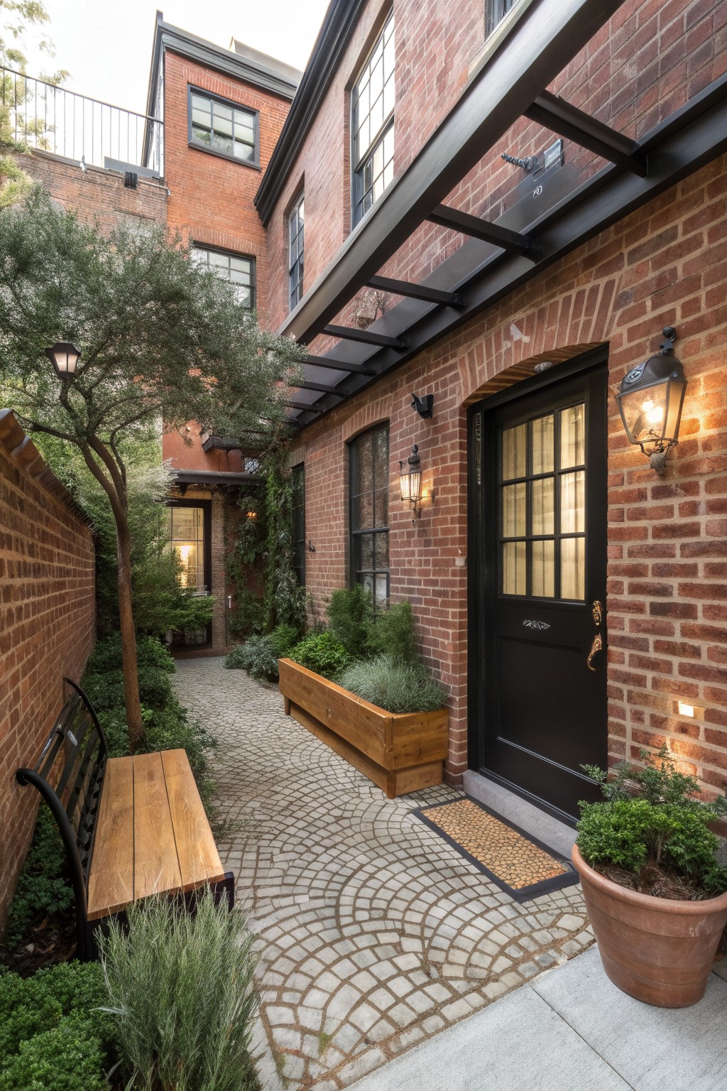 Narrow cobblestone pathway lined with plants, a wooden bench, and lanterns leading to a black door set in a red brick wall surrounded by brick buildings and greenery.