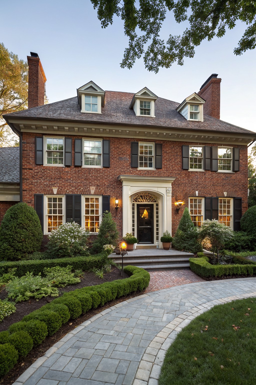 Two-story red brick house with dormer windows, black shutters, arched front door with lanterns, boxwood shrubs bordering a curved gray paver pathway and brick steps.