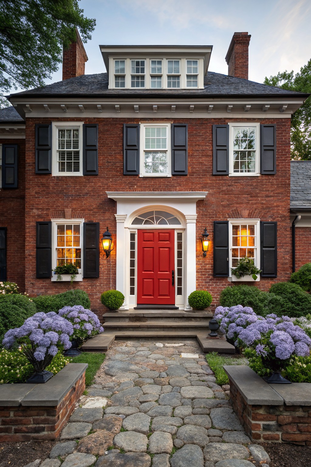Two-story red brick house with black shutters, white trim, arched entry porch, bright red front door, lanterns on sides, stone steps, cobblestone path, and purple hydrangea bushes in planters.