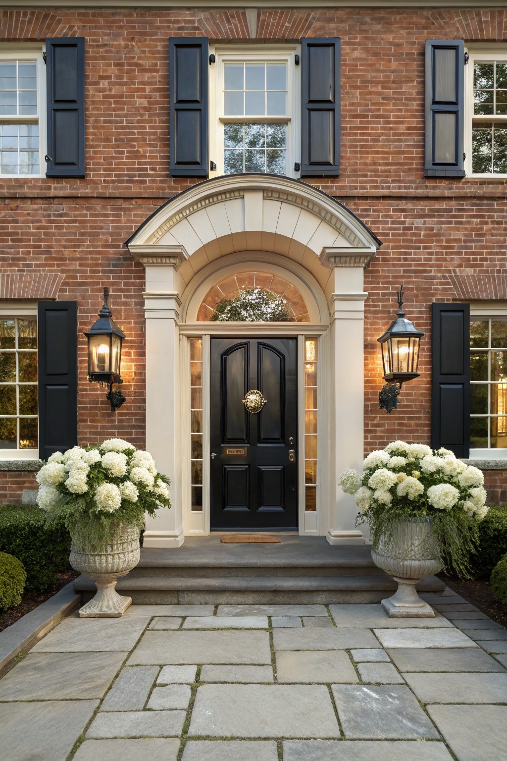 Red brick house facade featuring a centered black wood front door with brass knocker under a white arched surround, flanked by black lanterns and large white hydrangea arrangements in urns on stone steps, with black shutters on white-framed windows.