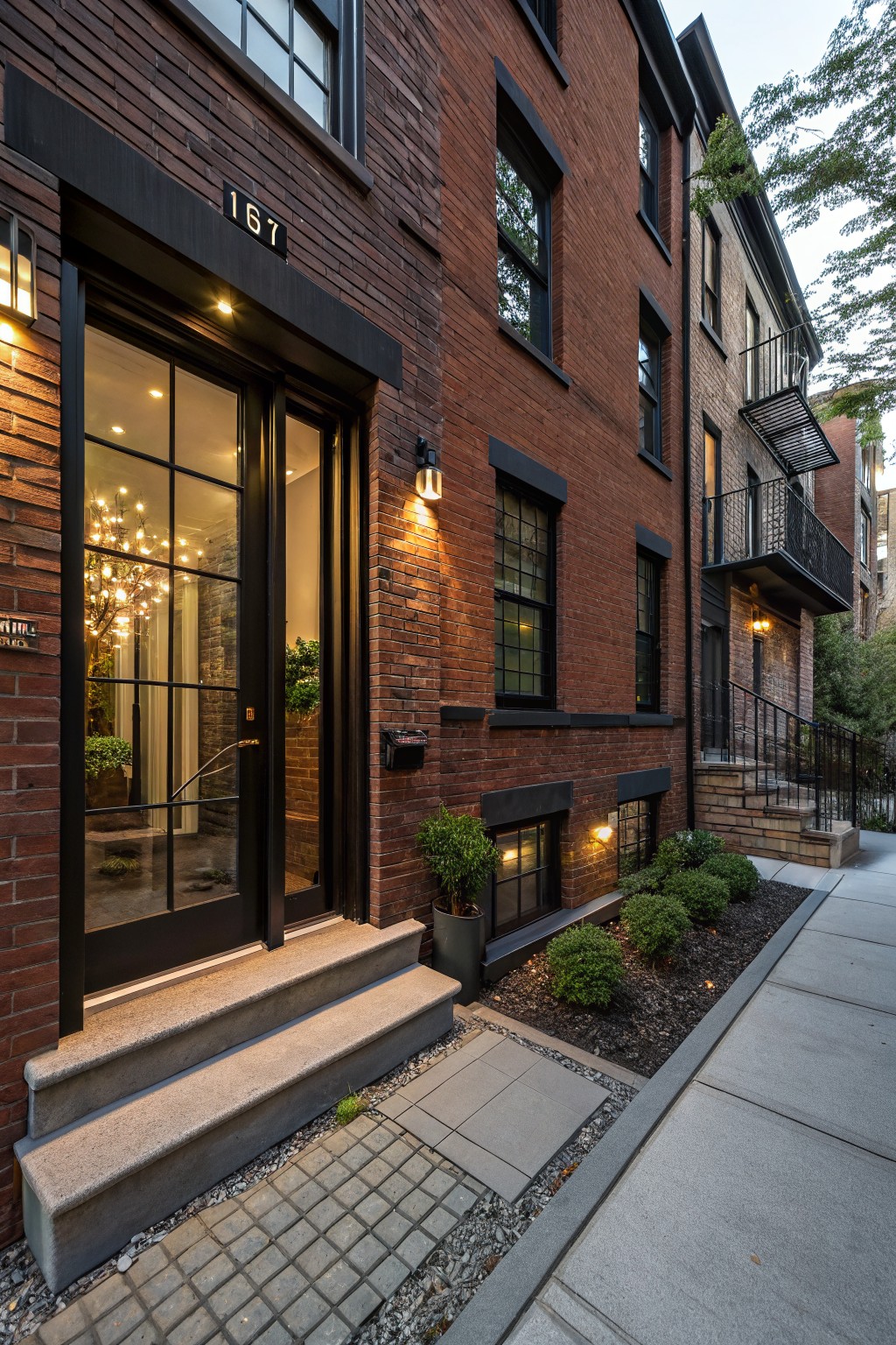 Red brick townhouse exterior with number 167 above black-framed glass double doors, flanked by lit lanterns, and low shrubs along concrete steps and sidewalk.