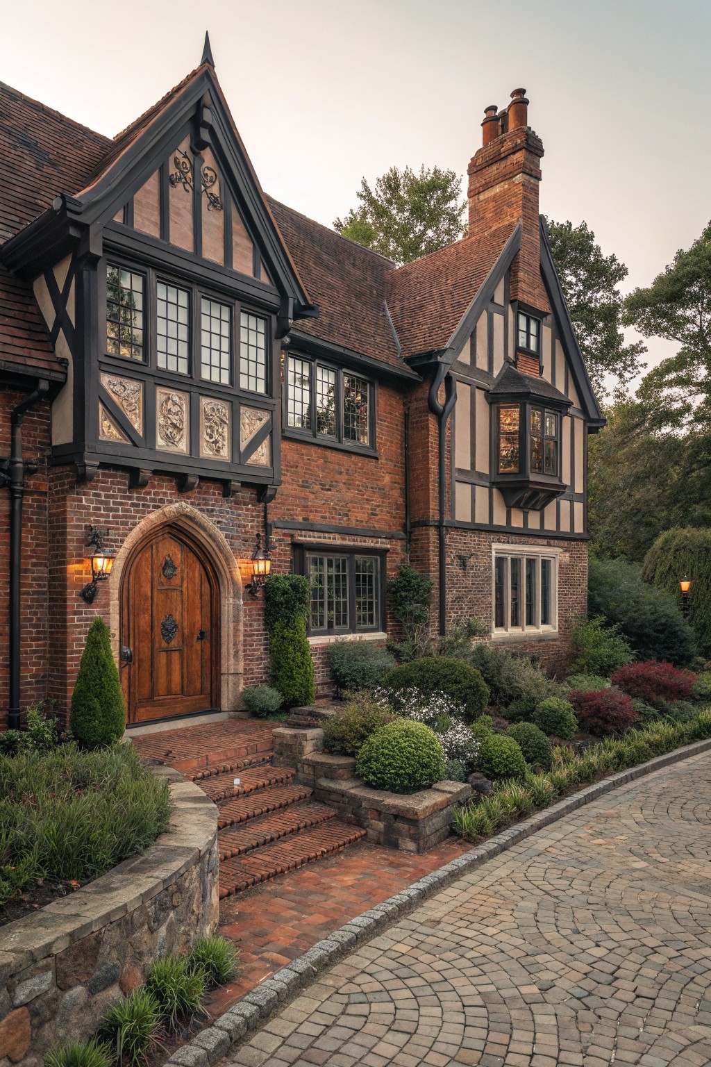 A two-story Tudor Revival house with red brick walls, black half-timbering, steeply pitched tile roofs, an arched wooden front door flanked by lanterns, brick steps, low stone walls, shrubs, and a curved cobblestone driveway.
