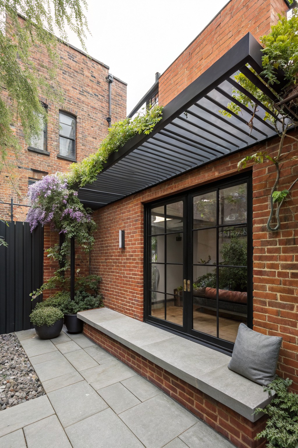 Red brick house wall with black metal pergola covered in green vines and purple flowers, concrete bench with gray cushion, potted plants, and gray paver patio with gravel edging.