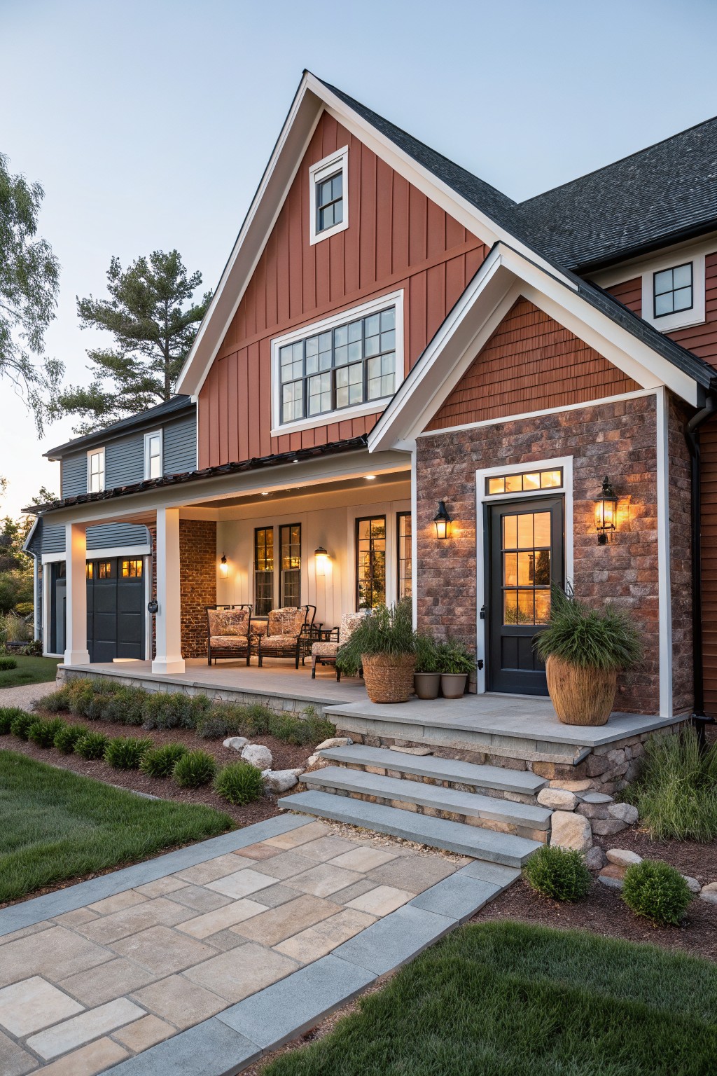 A two-story house exterior with red board-and-batten siding on the upper facade, brick base and entryway, covered front porch with wicker furniture and lanterns, stone steps, bluestone path, and low landscaping.