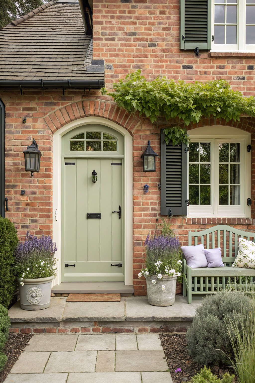 Red brick house facade with sage green arched front door, flanked by black lanterns, potted lavender plants, green bench with cushions, and climbing vines over the entry arch.