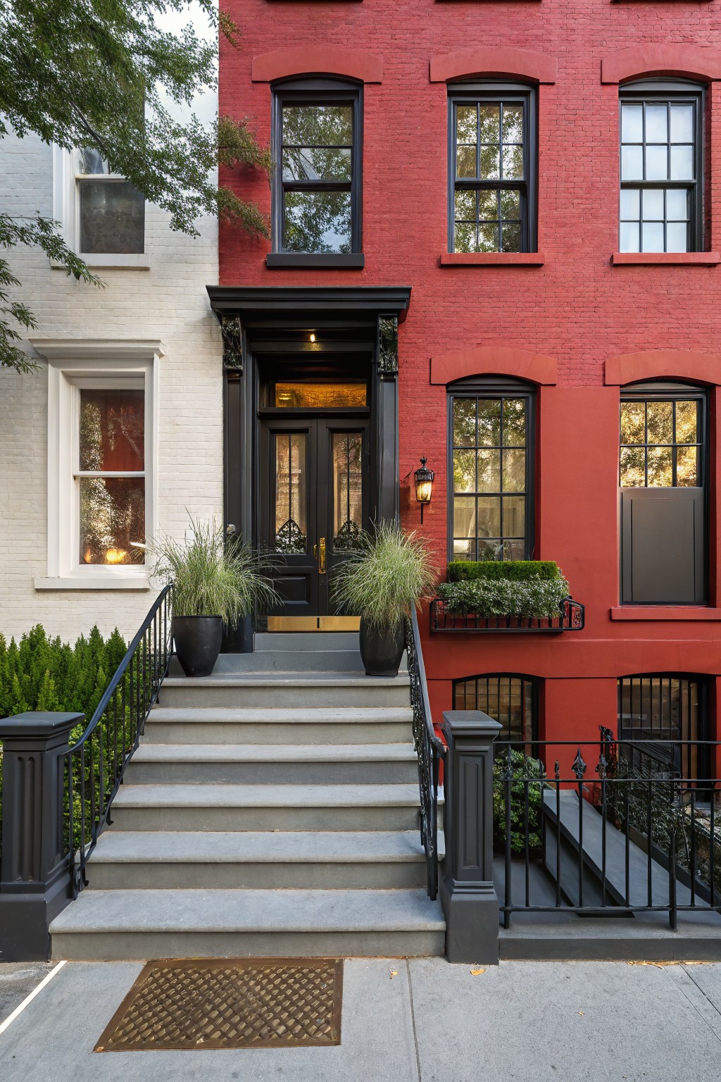 Red painted brick rowhouse with black window frames, double front doors, stone entry steps with black iron railings, potted grasses, and boxwood planter on windowsill next to white brick neighboring house.
