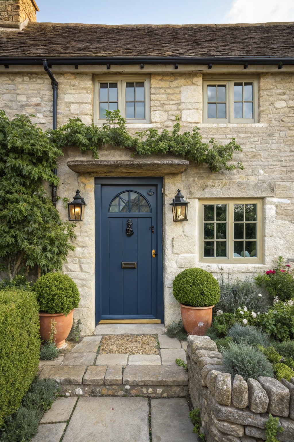 A two-story stone cottage exterior with beige stone walls, a navy blue arched wooden front door with brass knocker, cream-framed windows, black lanterns, climbing vines, potted boxwood topiaries, and a stone path.