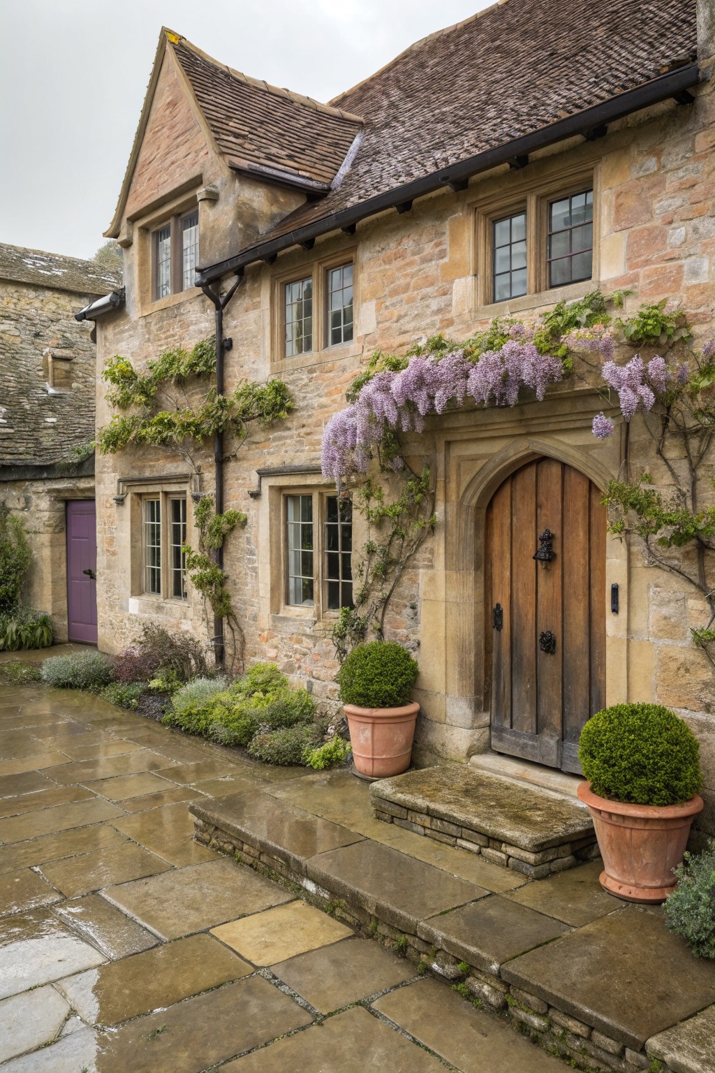 Stone house exterior featuring an arched wooden door partially covered by blooming purple wisteria vines, with climbing plants on the walls, potted topiary shrubs, and a wet stone pathway leading to steps.