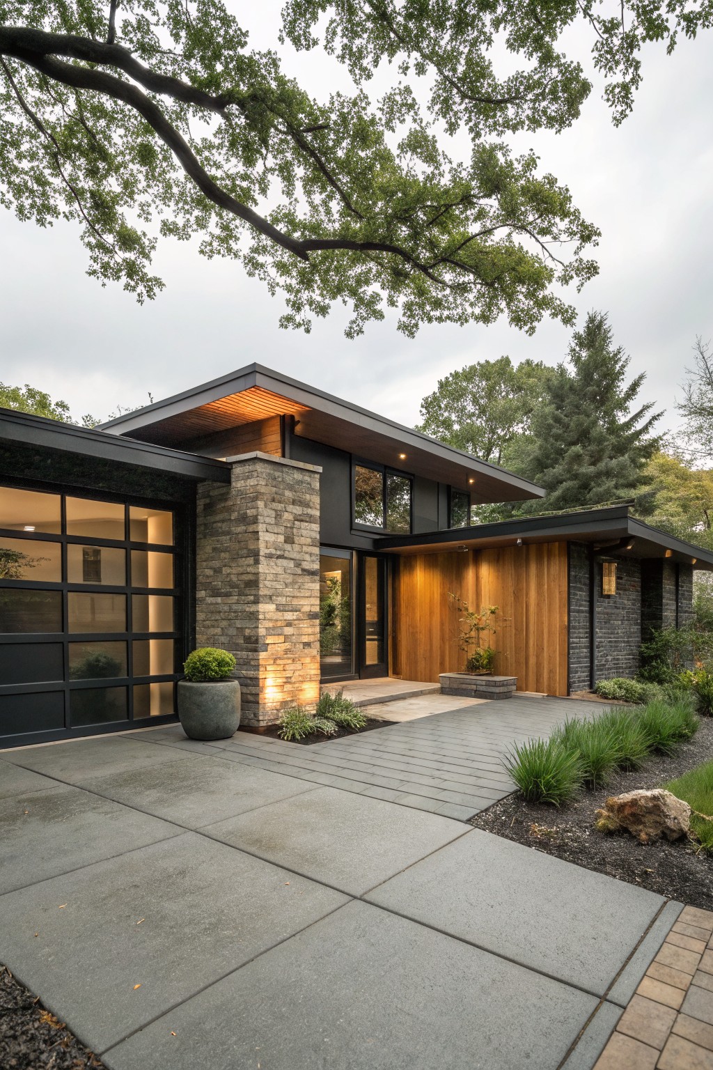 Modern house exterior with black glass garage door, tall stone pillar beside wood-paneled entry door, potted plants, paved driveway, and trees under cloudy sky.