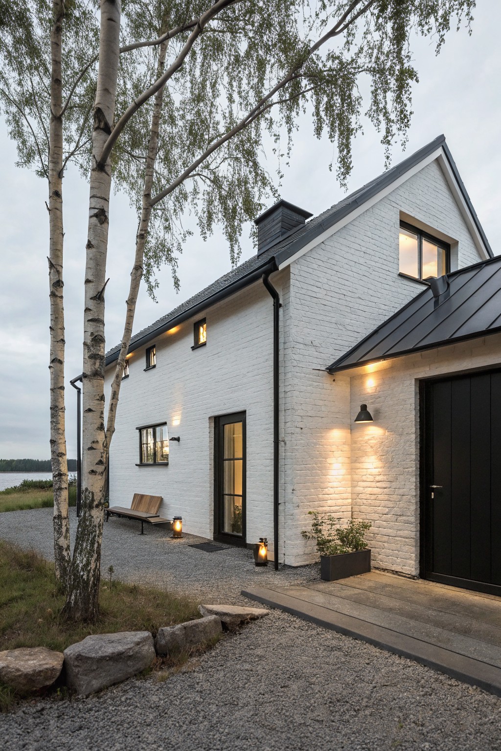 White painted brick house exterior with black metal roof, black garage door, lit lanterns, wooden bench, birch trees, gravel path and stone edging near a lake at dusk.