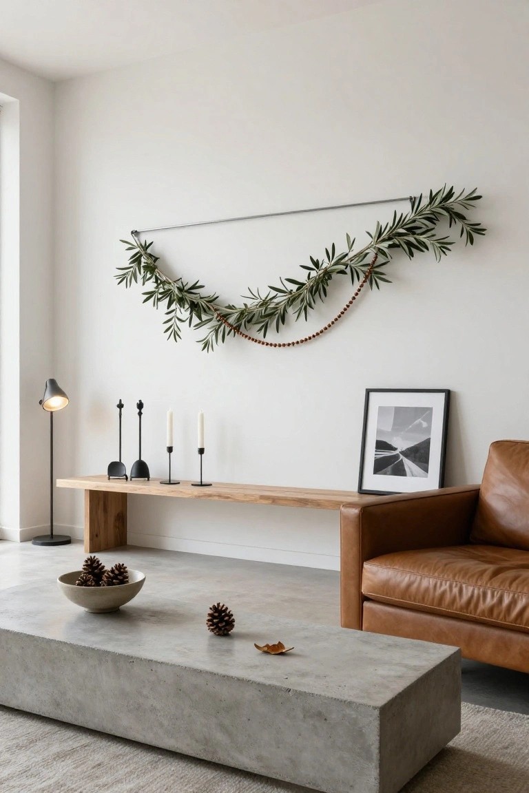 Modern living room corner featuring a light wood bench shelf with black candle holders and white candles, olive branch garland with wooden beads draped on a metal rod above, tan leather armchair, and concrete coffee table holding a bowl of pinecones.