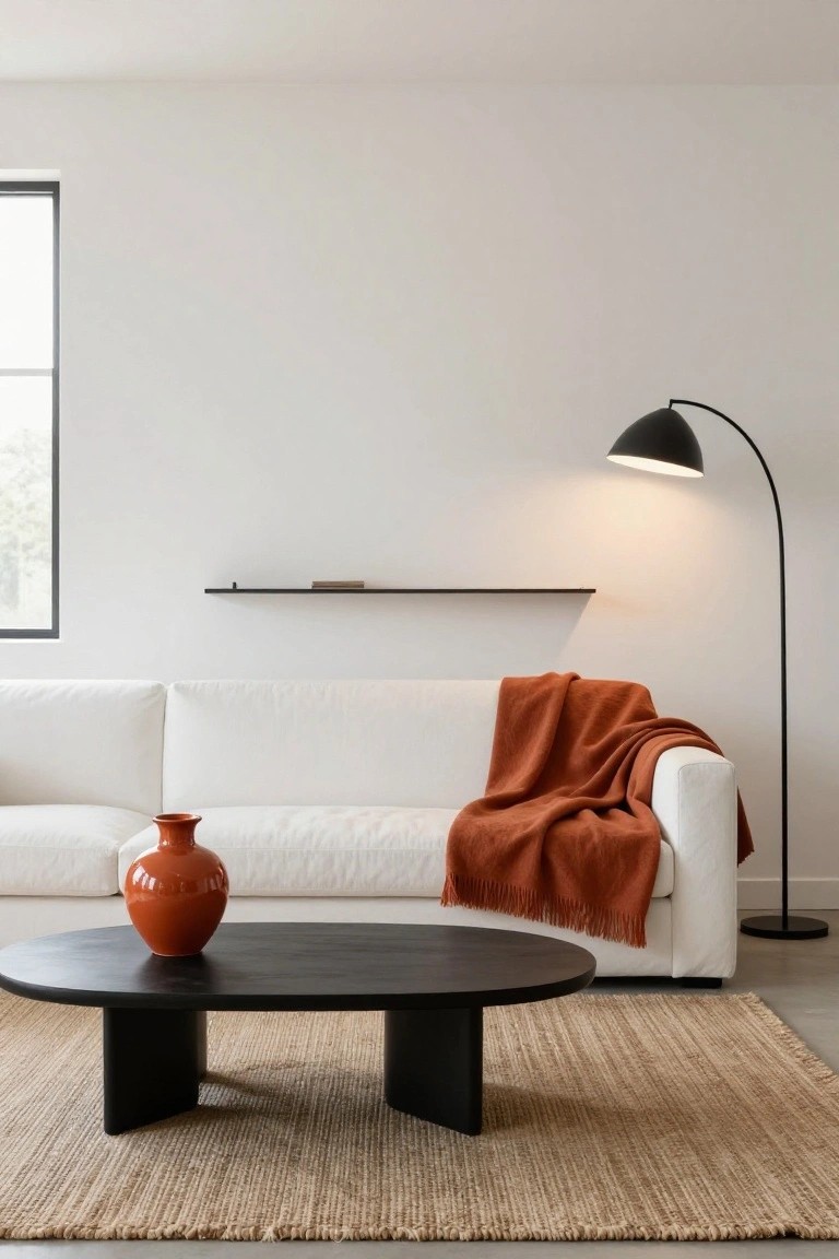 Minimalist living room with white sofa draped in orange blanket, black oval coffee table holding orange vase, seagrass rug, black arc floor lamp lit up, black wall shelf, and large window on white walls.