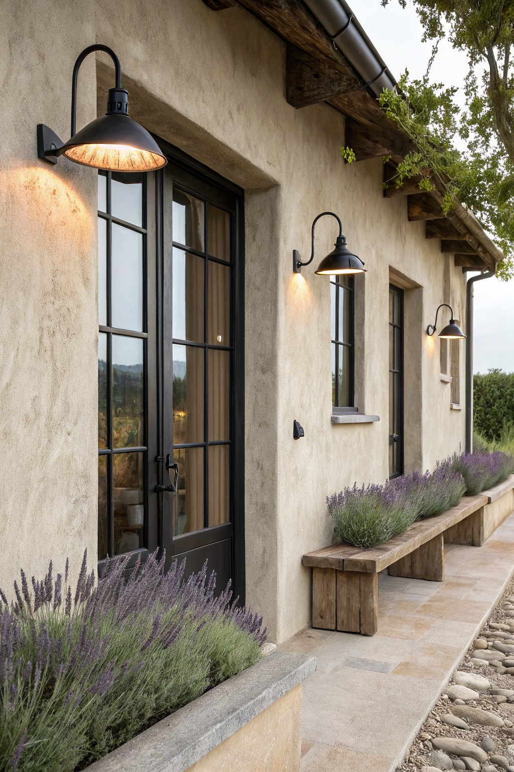 Beige stucco exterior wall of a house with exposed wood beams, black-framed glass doors and windows, black gooseneck wall lanterns emitting warm light, lavender plants in raised beds, wooden benches, and a stone path with gravel.