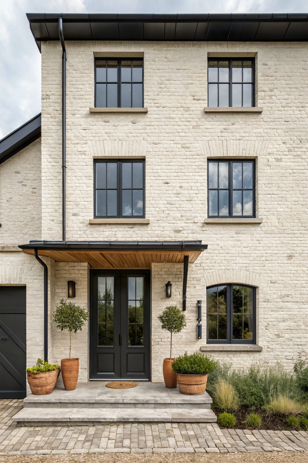 Two-story white brick house exterior with black-framed windows and double front doors under a wooden porch overhang, flanked by two black wall lanterns and potted plants on stone steps leading to a gravel path.