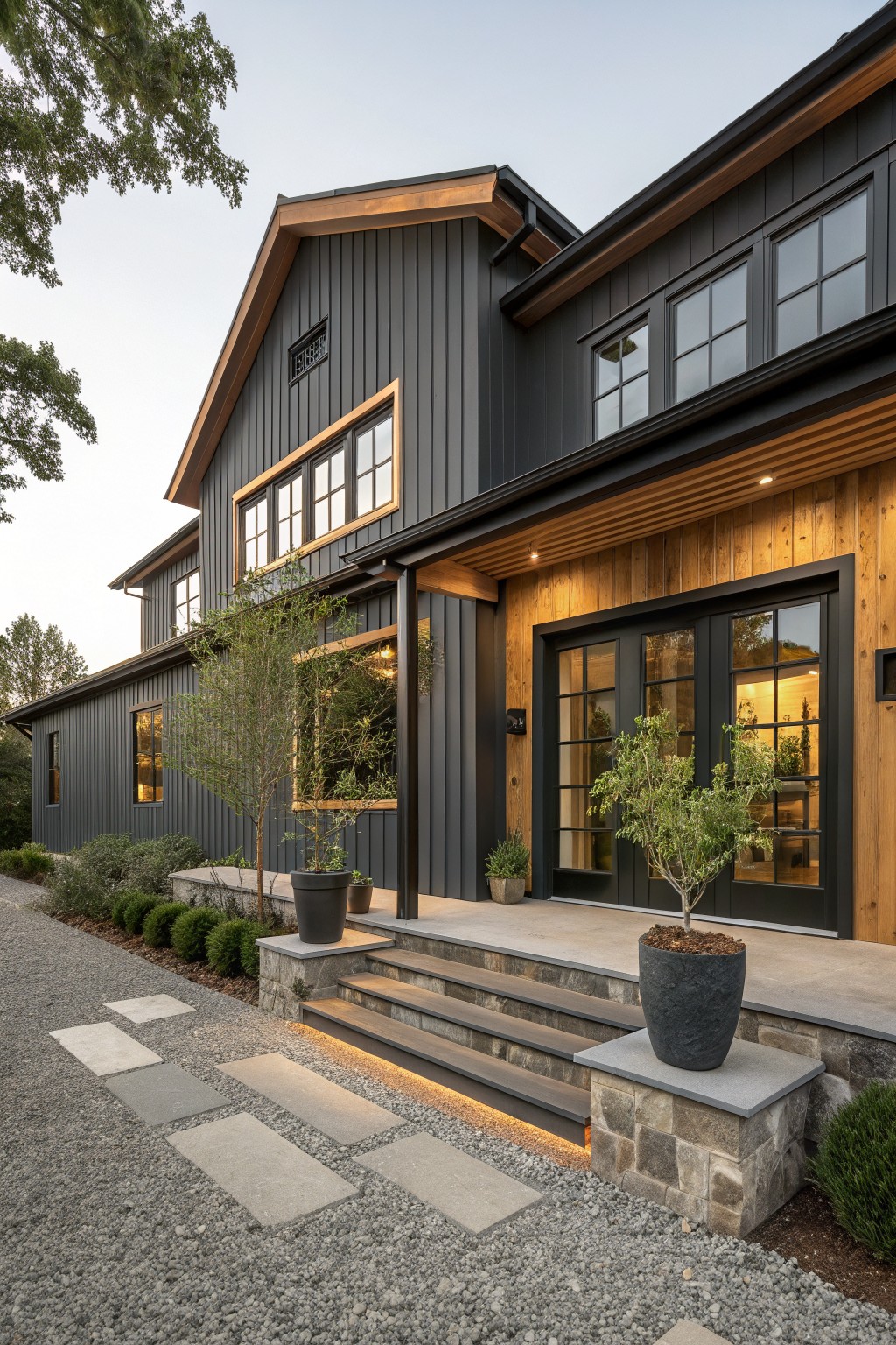 Two-story modern farmhouse exterior with dark gray board-and-batten siding, warm wood accents on the covered entry porch, black-framed windows, stone steps leading to glass doors, potted plants, and a gravel path edged with shrubs.