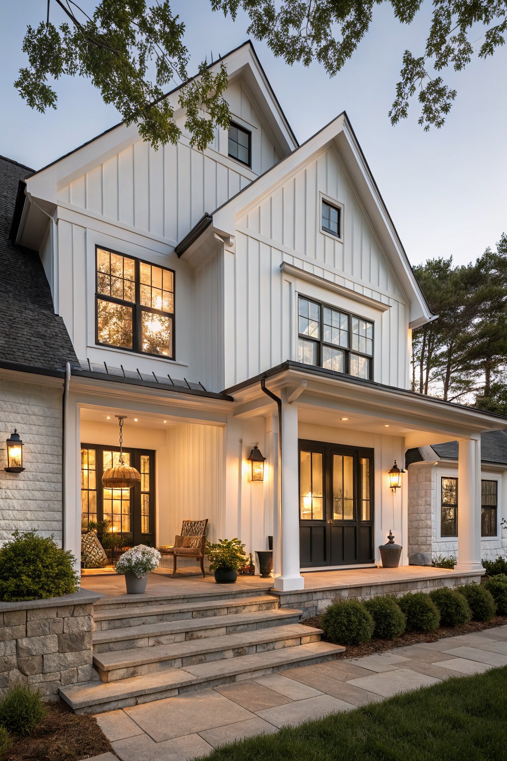 White board-and-batten sided modern farmhouse exterior with gabled roof, black windows and doors, covered porch with columns, stone steps, lanterns, potted plants, shrubs, and lawn at dusk.