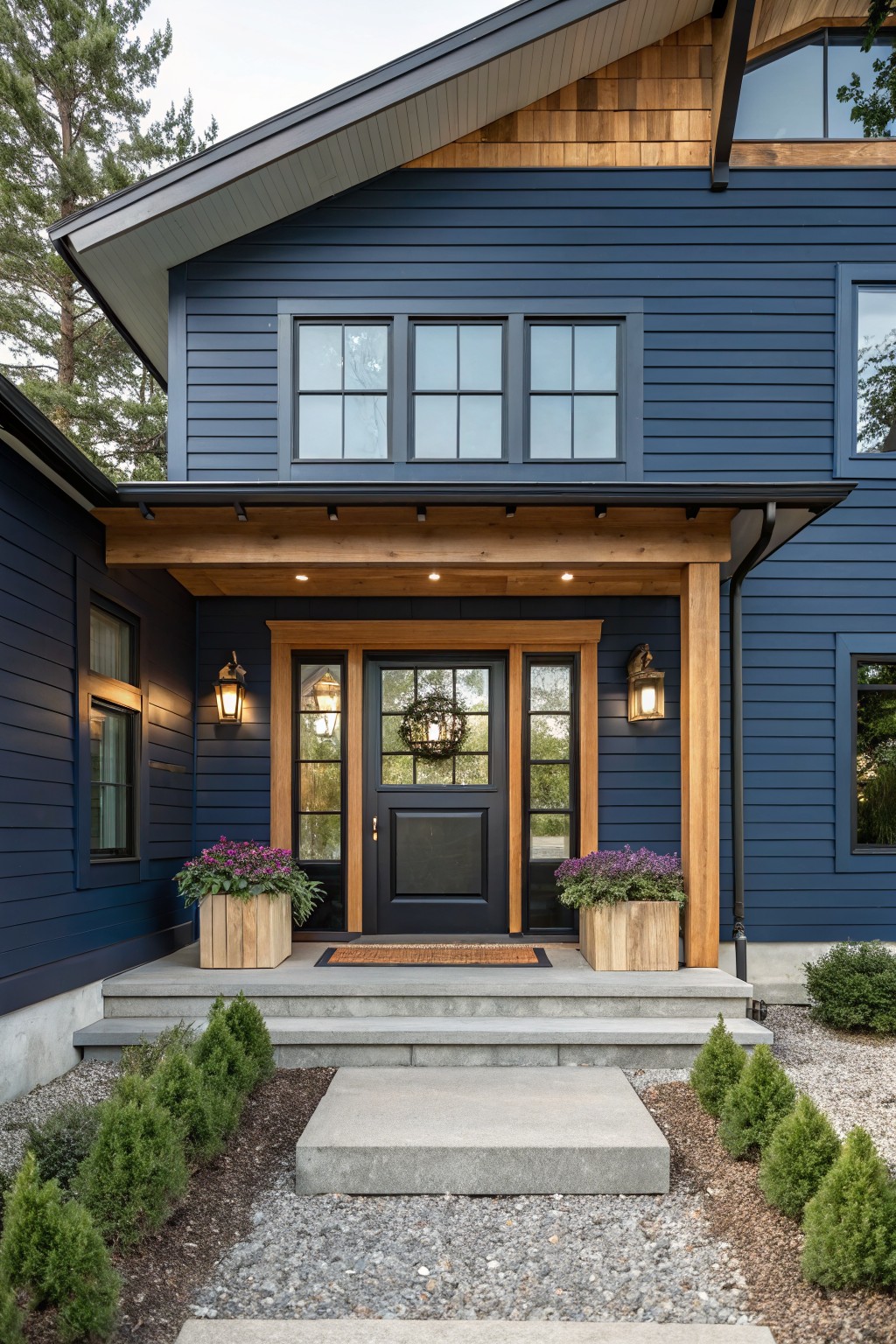 A two-story modern farmhouse with navy blue horizontal siding, wood-beamed covered entry porch, black-framed windows, potted purple flowers flanking a black front door, and boxwood shrubs along a concrete entry path amid gravel and pine trees.