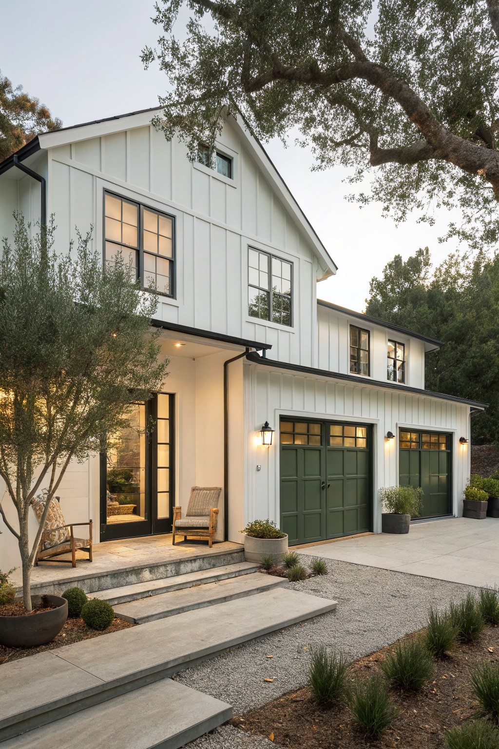 Two-story modern farmhouse house exterior with white shiplap siding, black-framed windows and doors, hunter green double garage doors, entry porch with lanterns and chairs under an olive tree, concrete steps, and gravel path with plants.