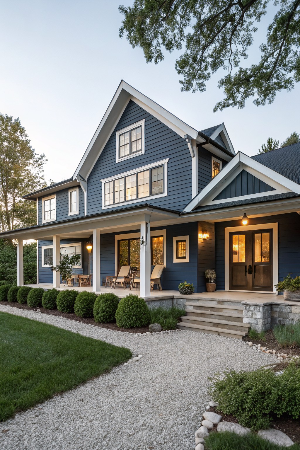 A modern farmhouse house exterior with navy blue clapboard siding, white trim, gabled roof, wraparound porch with columns, wooden double front doors, and landscaping including boxwood shrubs, gravel path, and lawn.
