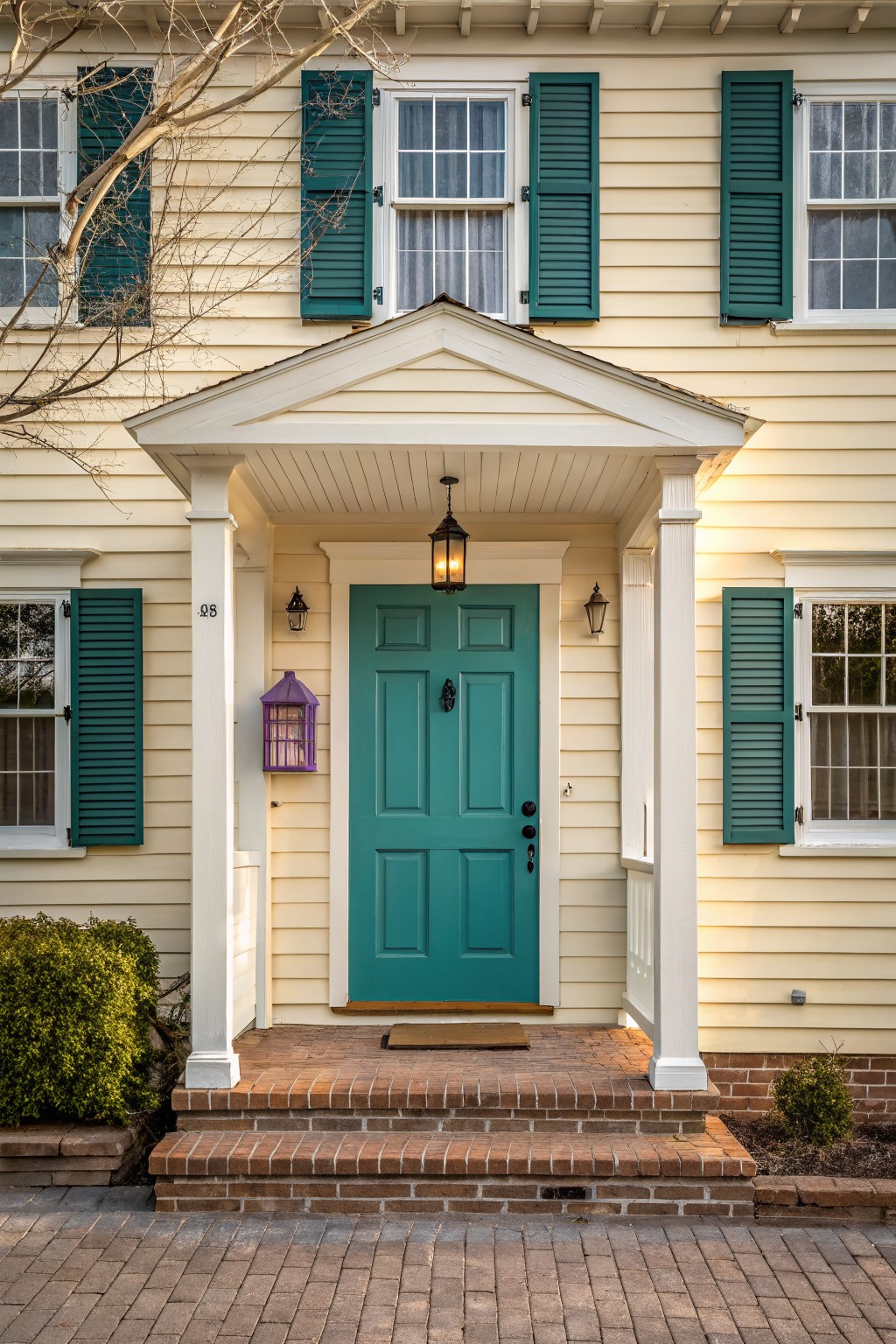 Front exterior of a two-story pale yellow house with teal front door, green shutters on windows, white porch supported by columns, lanterns, purple hanging lantern, brick entry steps, and paver walkway.