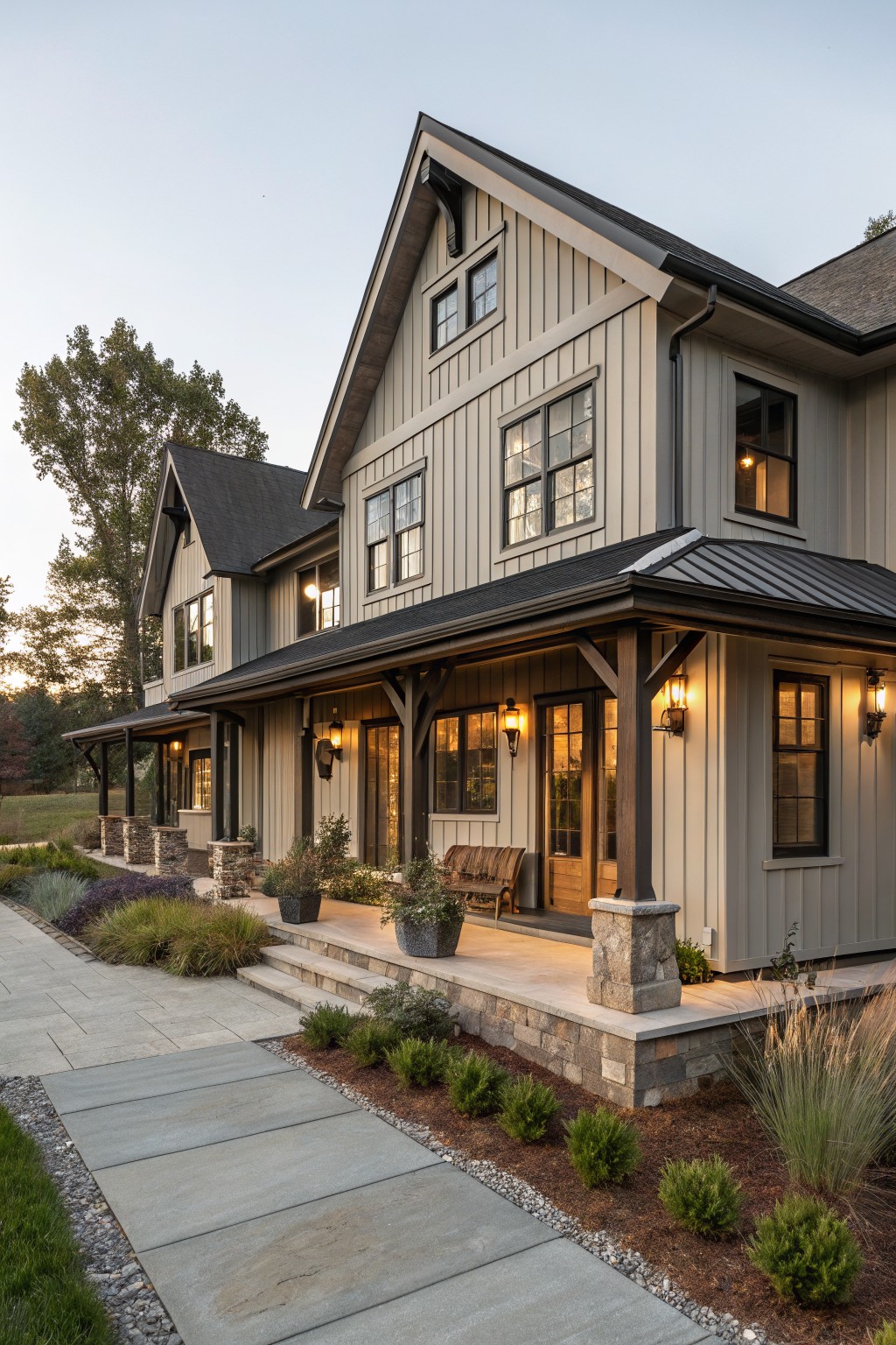 Side view of a two-story modern farmhouse with light gray vertical board and batten siding, black metal roof, covered porch with wood beams, stone steps, potted plants, and gravel path edged by shrubs at dusk.