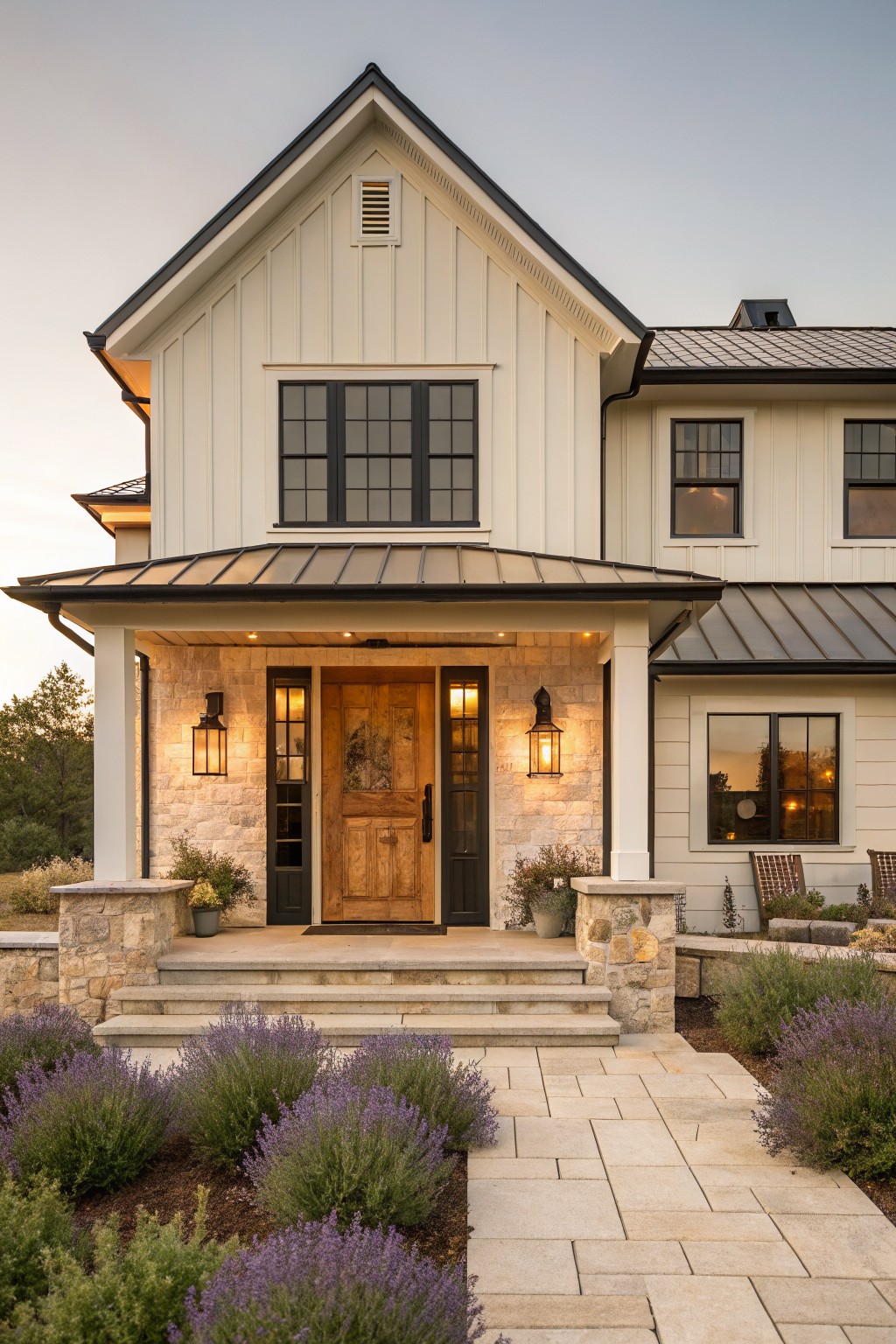 Front exterior of a modern farmhouse with white board-and-batten siding, black window frames, dark metal porch roof, wooden door flanked by lanterns, stone pillars and steps, and lavender plants along a stone pathway.