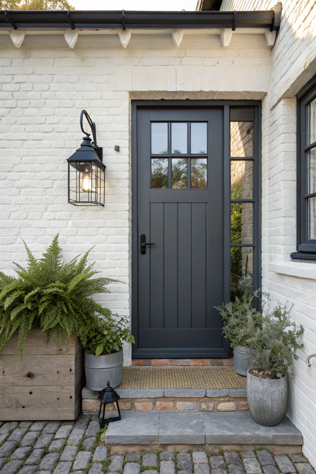White brick house exterior corner with dark gray paneled front door featuring glass upper panels, black wall lantern, potted ferns and shrubs, stone steps, and cobblestone path.