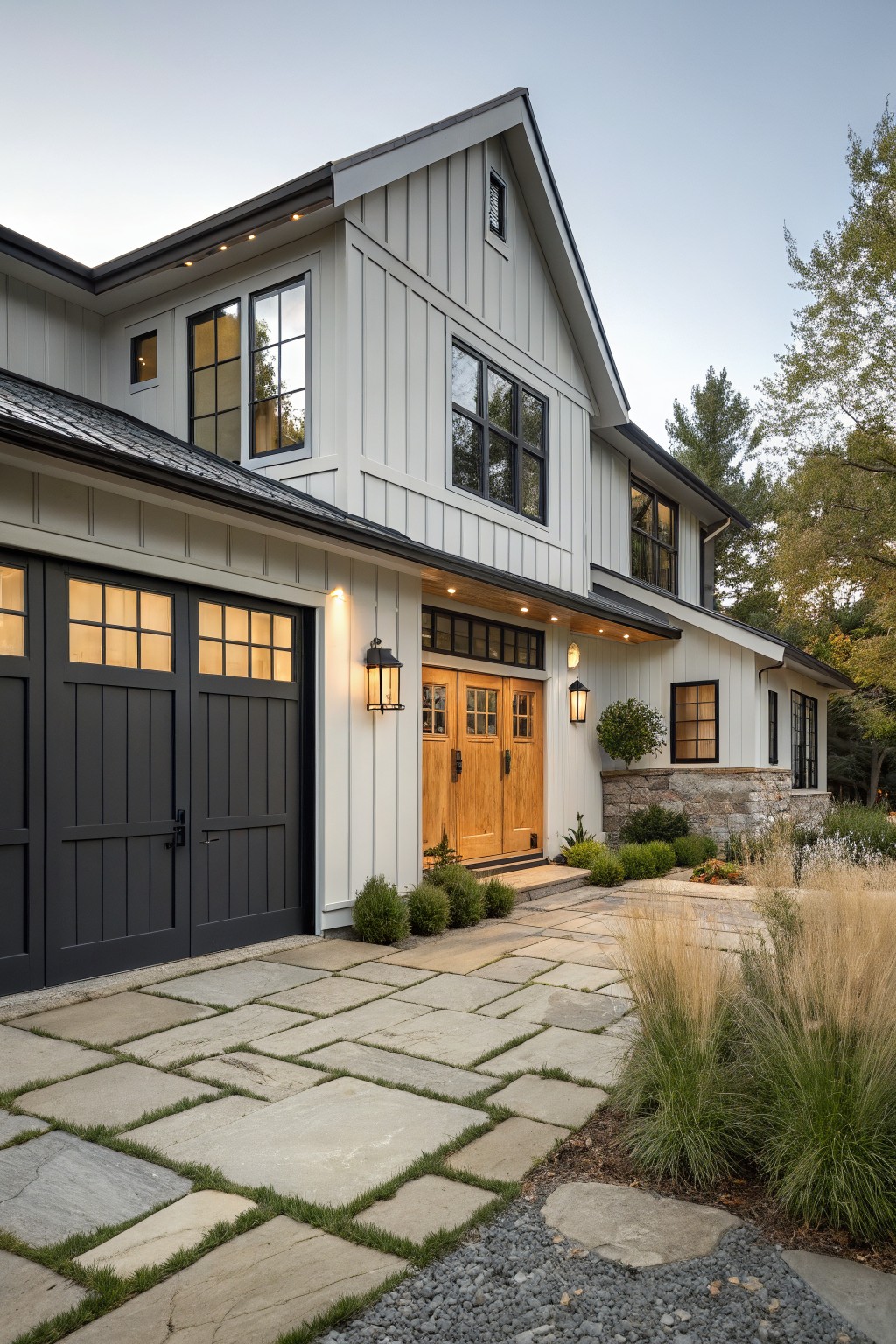 Modern farmhouse exterior featuring white board-and-batten siding, dark paneled garage doors, light-stained wooden double front doors with black hardware, wall lanterns, stone paver walkway, and low plantings.