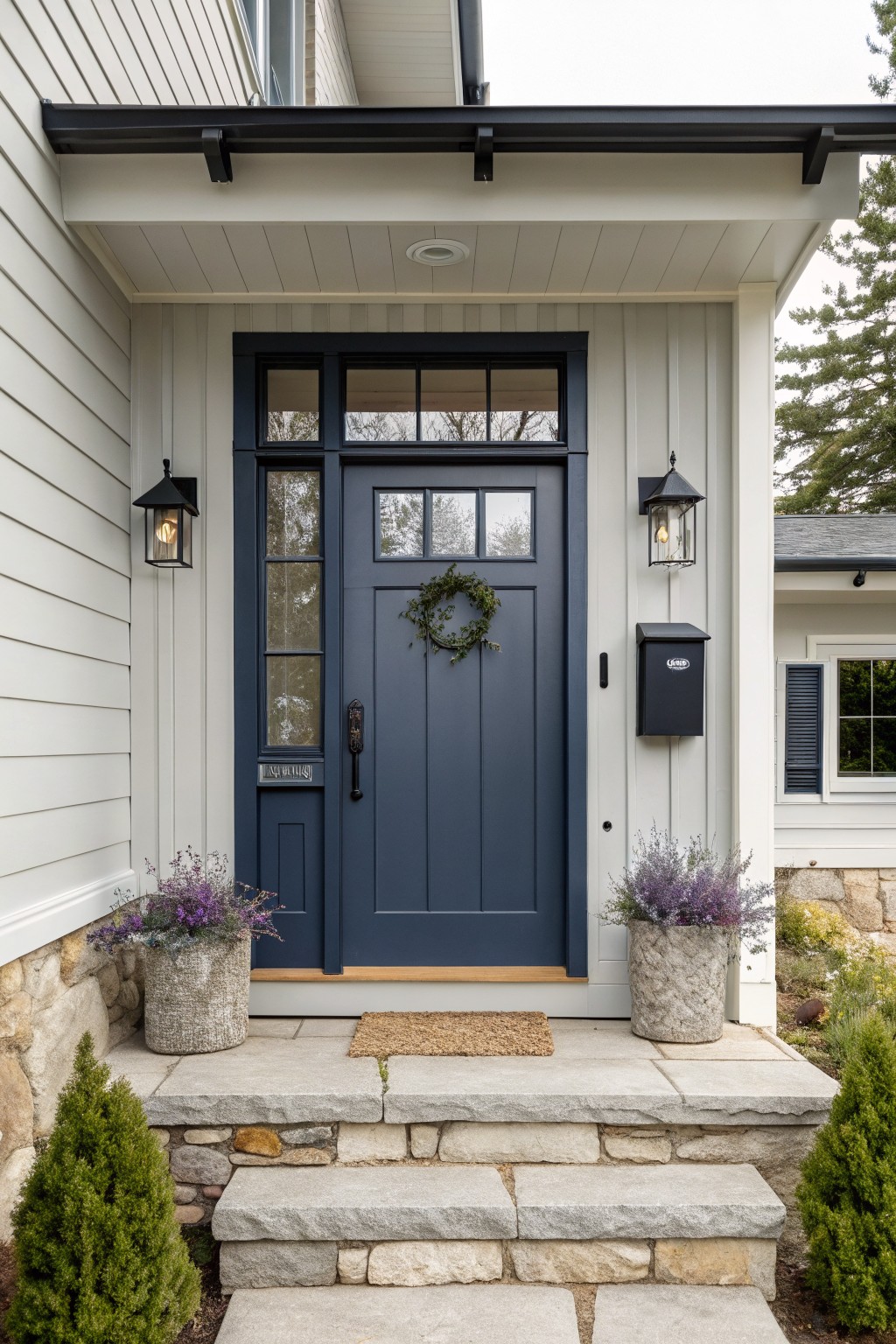 Modern farmhouse front entry with navy blue paneled door featuring glass windows, flanked by black lanterns, evergreen wreath, lavender plants in textured pots, doormat on porch, and stone steps with light shiplap siding.