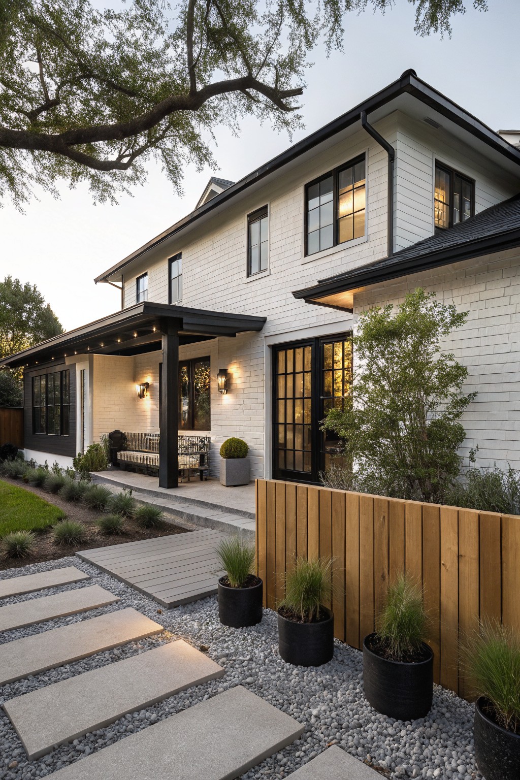 Side exterior of a white brick ranch house with black-trimmed covered porch, black-framed French doors, bench seating, concrete paver pathway through gravel and grasses, potted plants, and wooden fence.