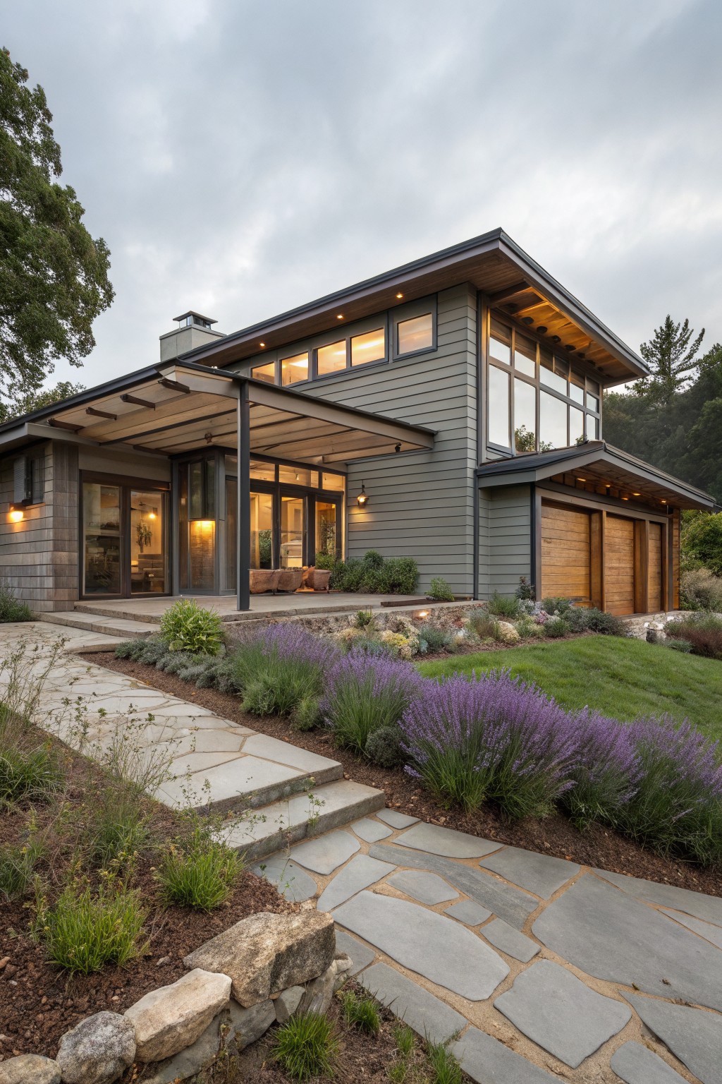 Gray modern ranch house exterior with overhanging roof covering a porch area with glass doors and metal supports next to wood garage doors, stone pathway with lavender plants leading up from landscaped beds, large windows, and surrounding trees under cloudy sky.